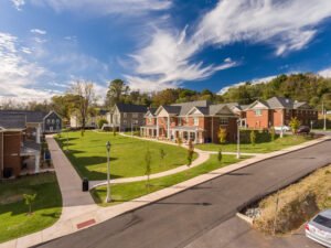 Low-altitude drone view of the Emory & Henry College student housing quad, highlighting manicured green lawns, curved concrete sidewalks, traditional lamp posts, and red brick residential buildings under a bright blue sky with wispy clouds.