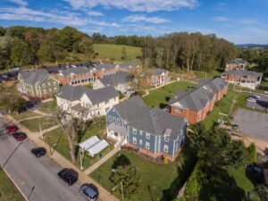 High-angle aerial drone view of Emory & Henry College student village housing, highlighting blue, yellow, and green clapboard residences alongside brick dormitory halls, paved streets, and parking areas against rolling green hills.