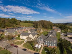 Aerial drone view of the Emory & Henry College student housing village, showing multi-story residential buildings with blue, yellow, and green siding, sprawling parking areas, and rolling green hills under a bright blue sky.