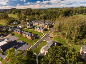 Aerial drone overview of the Emory & Henry College student village housing development featuring brick multi-unit residential halls, green lawns, parking lots, and surrounding rolling wooded hills under a blue sky.