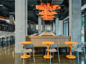 Straight-on view of vibrant orange geometric acoustic ceiling baffles, raw concrete pillars, and booth seating at UTK Cumberland Food Hall in Knoxville, TN.