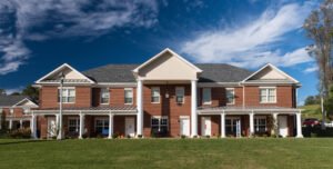 Straight-on daylight exterior architectural view of a two-story red brick student residential complex at Emory & Henry College, showcasing a central pediment portico, large white columns, metal porch roofs, white doorways, and a manicured green lawn under blue skies and wispy clouds.