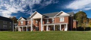 Low-angle exterior architectural shot of a two-story red brick student residence building at Emory & Henry College, showcasing white classical columns, standing-seam porch roofs, gabled rooflines, and a grassy lawn under a deep blue sky with scattered clouds.