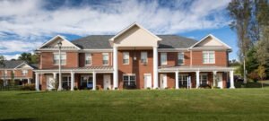 Straight-on daylight exterior architectural view of a two-story red brick student residence hall at Emory & Henry College, featuring a symmetrical facade, prominent gabled portico, white Tuscan columns, front porch walkways, and a manicured green lawn under blue skies.