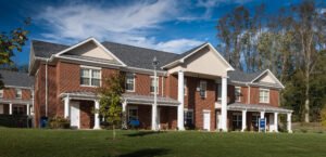 Angled exterior view of a brick student residential hall at Emory & Henry College, featuring a prominent gabled portico, white neoclassical columns, standing-seam porch roofs, and a green front lawn under a blue sky.