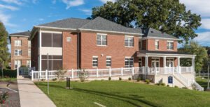 Angled exterior daytime view of the Emory & Henry Village Community Center, featuring red brick construction, large divided-lite windows, an accessible walkway with white balustrades, and a covered porch supported by white columns against a green lawn and blue sky.
