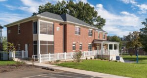 Three-quarter exterior architectural photograph of the Emory & Henry Village Community Center showing the red brick building corner, expansive floor-to-ceiling windows, white accessible ramp balustrades, and a covered porch on a sunny day with blue skies and clouds.