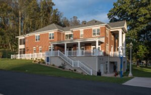 Front three-quarter exterior view of the Emory & Henry Village Community Center, showing red brick facade, illuminated portico porch, white columns and railings, concrete access staircase, and wooded hillside backdrop under late-afternoon light.