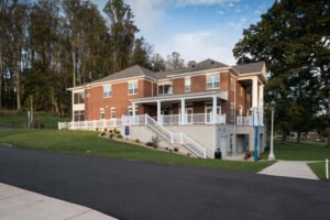 Exterior view of the Emory & Henry Village Community Center, featuring a red brick multi-story building with white porch railings, classical columns, concrete entrance stairs, and a walk-out lower level bordered by trees.
