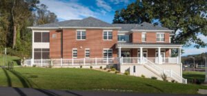 Straight-on daylight exterior architectural view of the Emory & Henry Village Community Center, featuring red brick masonry, a covered columned porch, white railings, concrete access stairs, ADA ramps, and lush green lawn against blue sky.