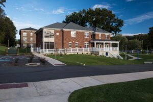 Wide daylight exterior view of the Emory & Henry Village Community Center, featuring red brick masonry, accessible ramp pathways with white railings, a covered pillared porch, and landscaped campus grounds beneath a blue sky.