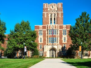 Architectural exterior photograph of Ayres Hall central brick clock tower, stone arch entrance, and American flag at UTK in Knoxville, TN.