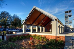 Low-angle exterior view of the University of Tennessee Band Complex pavilion showcasing the expansive gabled roof with wood plank soffit, heavy-duty industrial ceiling fan, brick planter wall, and steel observation tower under a blue sky.