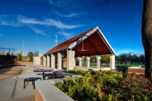 Angled exterior view of the open-air athletic pavilion at the University of Tennessee Knoxville, showcasing brick retaining planters, concrete seating plaza, stone support piers, and athletic turf fields under a vibrant blue sky.