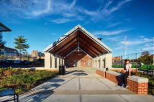Symmetrical exterior view of the modern open-air pavilion at the University of Tennessee Knoxville, featuring an expansive gabled timber roof, concrete walkway, brick planters, and a student seated on a brick wall beneath a clear blue sky.