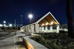 Dramatic night photograph of the University of Tennessee Band and Intramural Complex pavilion, featuring illuminated wood soffit, stone support columns, landscaped brick planter, patio seating, and bright stadium starburst lights under a dark night sky.