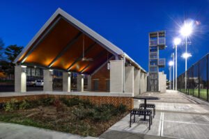 Wide-angle evening photograph of the UTK Band and Intramural Complex featuring a lit gabled pavilion, landscaped brick retaining wall, outdoor table, metal observation tower, and bright athletic field lights under a blue twilight sky.