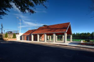 Wide daytime exterior of the UTK Band and Intramural Complex open-air pavilion featuring red textured gabled roof shingles, stone pillars, brick service building, and green athletic turf field under a clear blue sky.