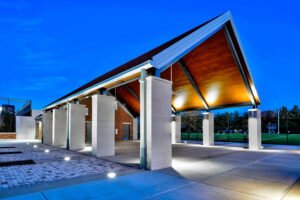 Twilight exterior photograph of the open-air pavilion at the University of Tennessee Knoxville, showcasing illuminated wood ceiling planks, concrete pillars, and paver plaza against a blue evening sky.