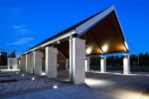 Twilight architectural view of the open-air pavilion at the University of Tennessee Knoxville, showcasing illuminated wood ceiling decking, square concrete support columns, exterior uplighting, and brick plaza pavers under a deep blue sky.