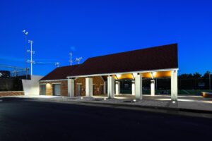 Wide twilight exterior view of the University of Tennessee Band and Intramural Complex pavilion and brick utility facility, featuring illuminated soffit lighting, square concrete columns, and paved parking drive under a deep blue dusk sky.