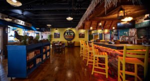 Panoramic commercial interior photograph of Over Easy Al’s Breakfast dining room featuring a stacked stone bar under a thatched tiki roof, bright yellow barstools, and rustic wood floors by Dwayne Hickson, Graphic Productions in Sevierville, TN.