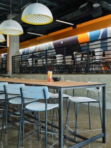 Modern high-top communal dining table, dome pendant fixtures, metal railing, and colorful wall mural at UTK Cumberland Food Hall in Knoxville, TN.