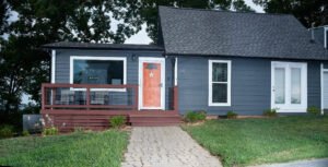 Exterior architectural photograph of a modern blue Douglas Lake Airbnb rental cottage with a coral front door, stone walkway, front porch deck, and lush lawn by Dwayne Hickson, Graphic Productions in Dandridge, TN.