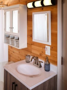 Cabin bathroom vanity with brushed nickel faucet, framed wall mirror, overhead lighting, and white hanging storage cabinet on pine tongue-and-groove walls.