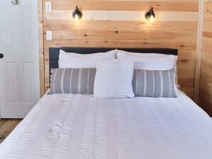 Straight-on view of a neatly dressed queen bed with white textured comforter, striped accent pillows, dark headboard, and dual warm wall sconces on pine shiplap.