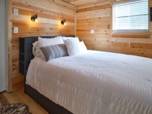 Rustic cabin bedroom with natural pine wood plank walls, black headboard with built-in shelves, made bed with white bedding, and warm wall sconces.