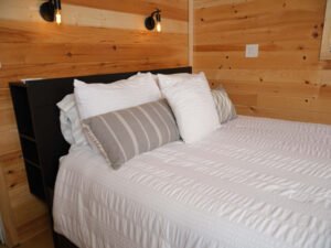 Cozy cabin bedroom with natural wood shiplap walls, wall-mounted reading sconces, and a made bed with white and gray linens.