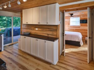 Modern white shaker cabinetry with dark stone countertop, wood backsplash, and view into an adjacent pine bedroom at a Douglas Lake cabin.