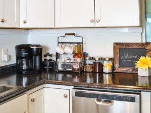 Fully stocked guest coffee bar featuring a Keurig coffee maker, French press, mug rack, syrups, and a rustic chalkboard welcome sign on dark kitchen countertops.