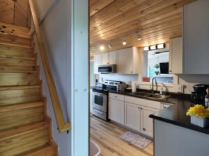 Modern cabin kitchen with stainless steel appliances, pass-through window to living area, and adjacent pine wooden staircase.