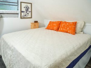 Loft bedroom under a sloped ceiling featuring a made bed with an off-white quilt, bright orange geometric accent pillows, and framed dog wall art.